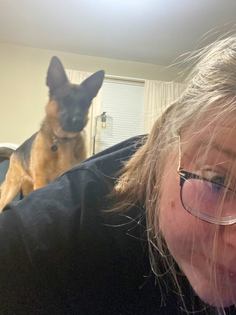 Close-up of a woman lying on the floor mid-workout, with a German shepherd puppy standing on her back and staring into the camera.