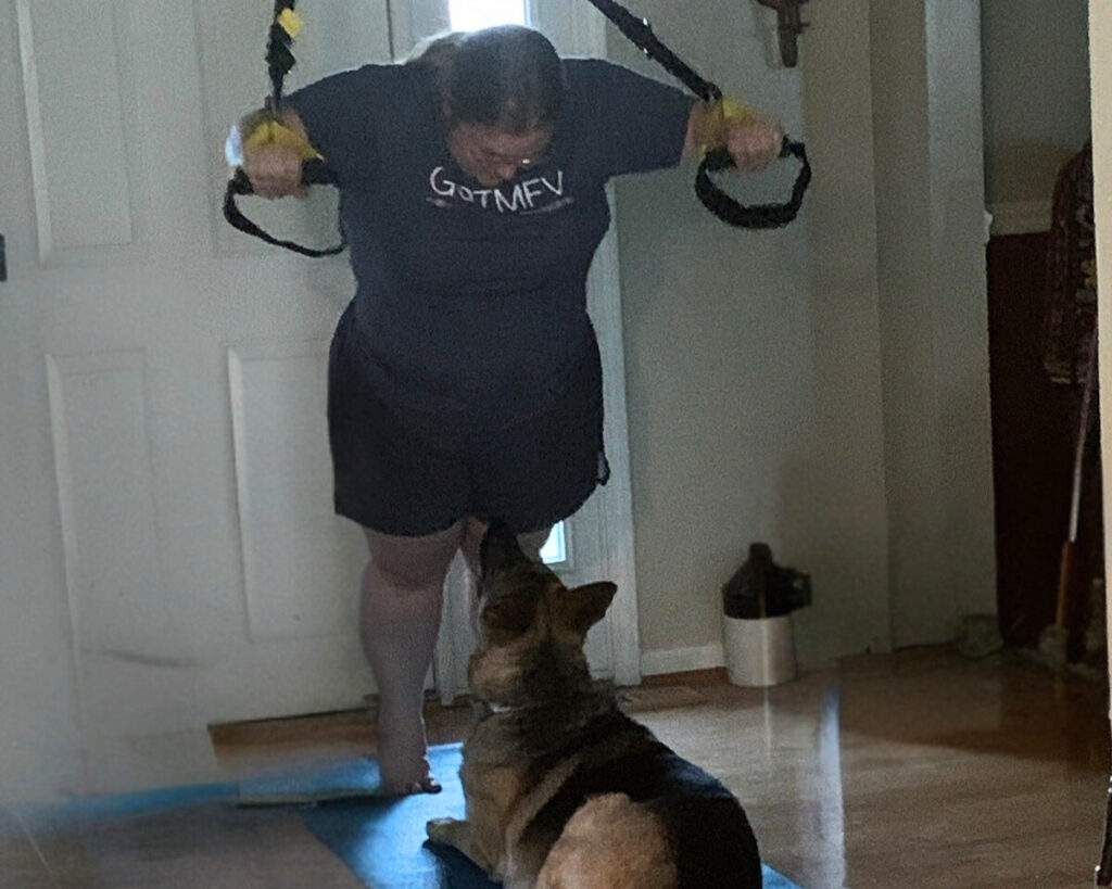 A woman using TRX straps for a home workout while her German shepherd sits directly in front of her on a yoga mat, looking up at her.