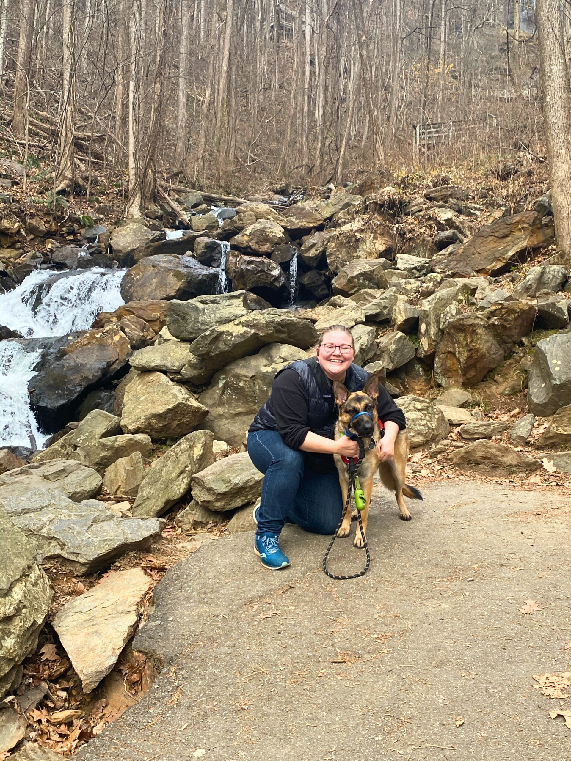Woman kneeling beside a German shepherd in front of a rocky waterfall on a forest hiking trail