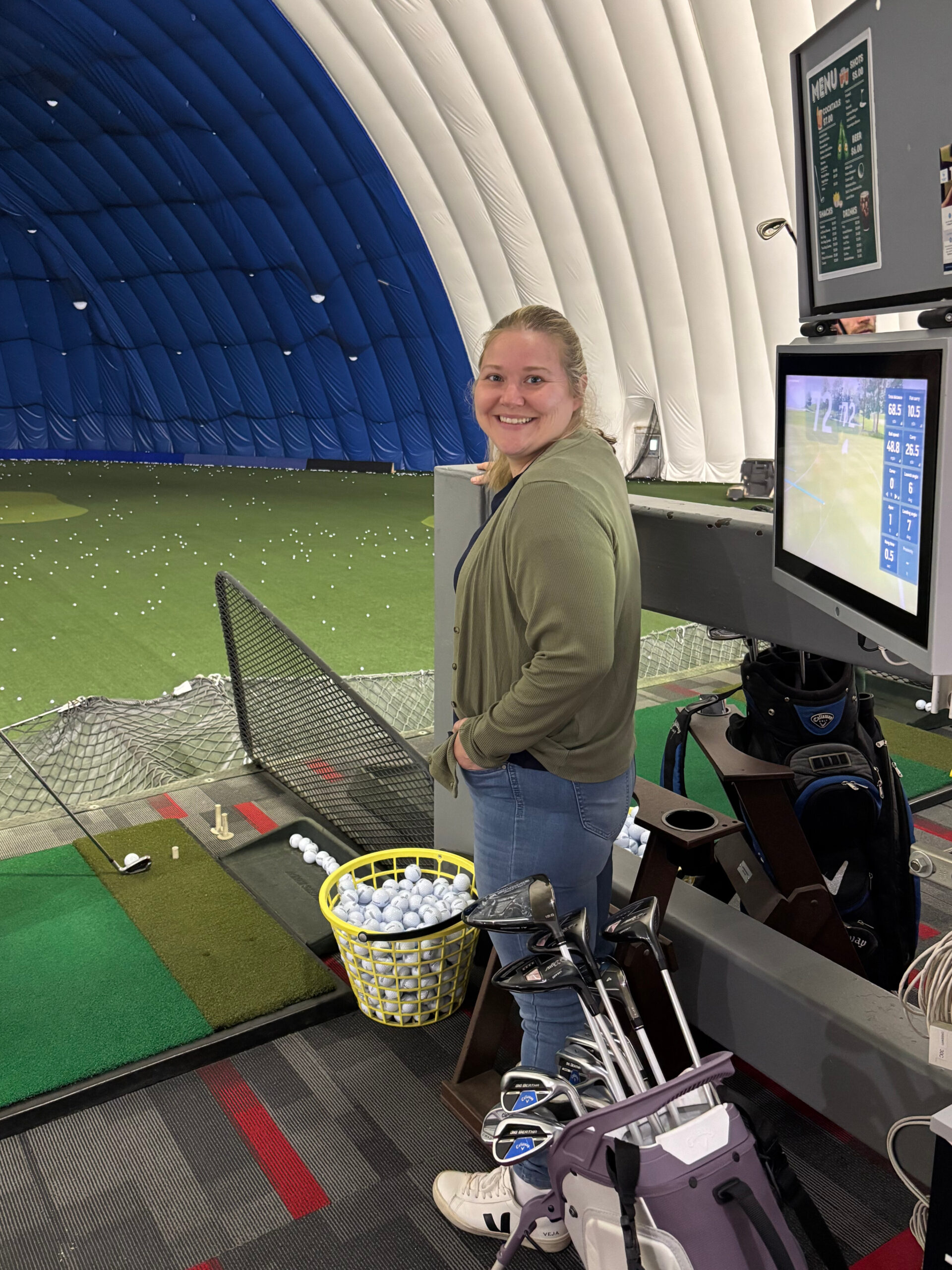 Woman smiling at an indoor driving range beside a simulator, helping her niece practice golf