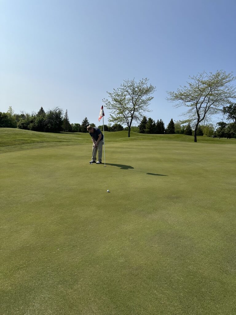 A golfer lines up a short putt with a mallet putter on a green with trees and a checkered flag in the background.